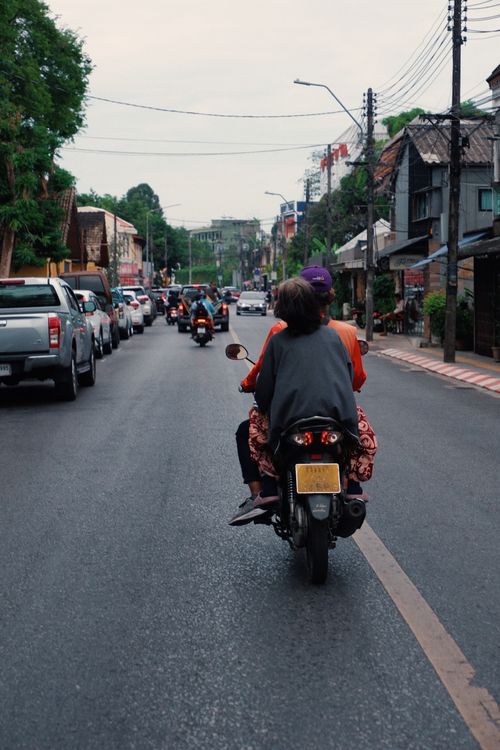 Free Two people riding a motorbike down a busy city street lined with cars and buildings. Stock Photo