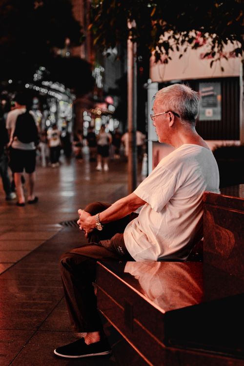 Free A senior man sitting on a bench in a lively city street illuminated by evening lights. Stock Photo