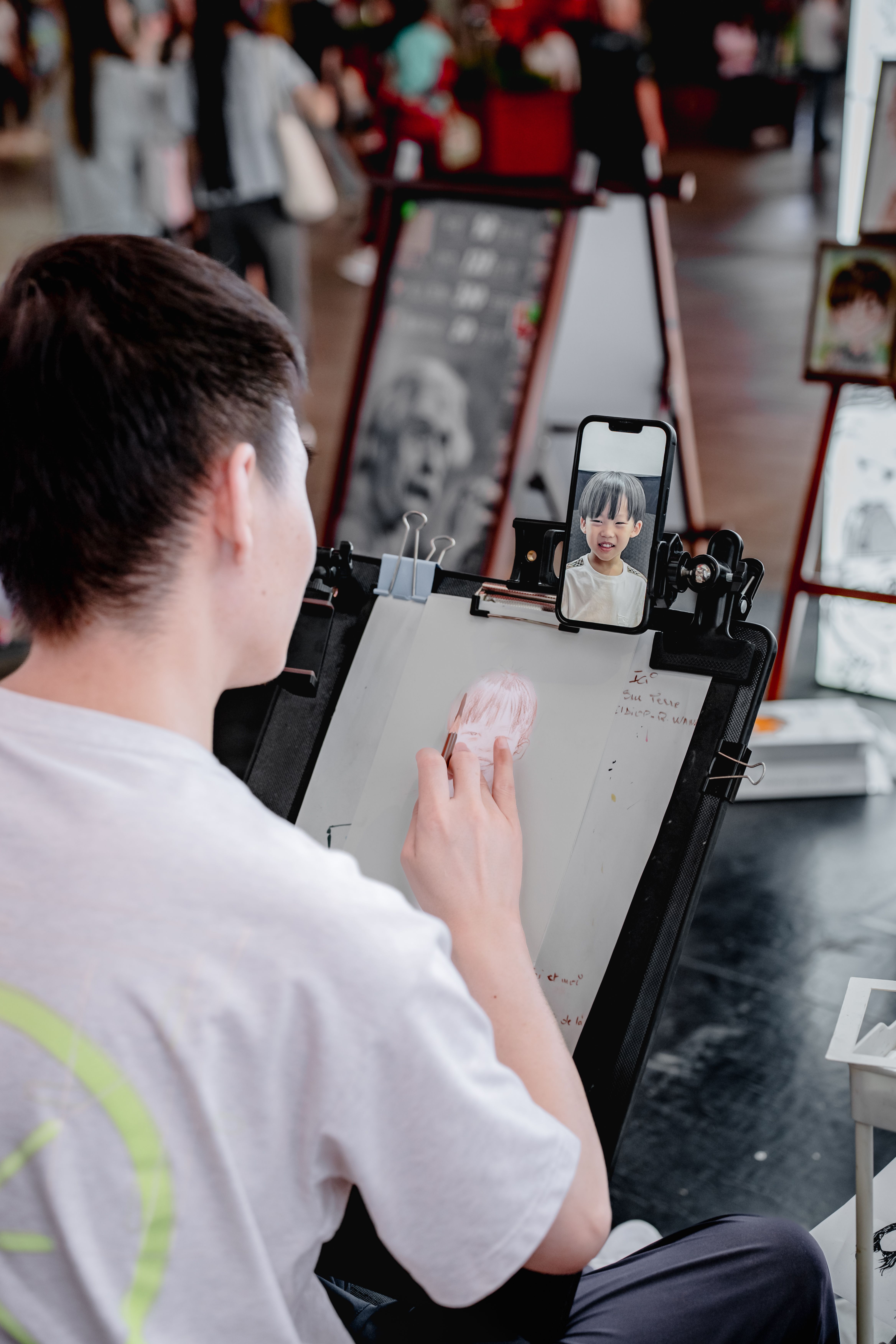 An artist draws a portrait using a smartphone reference in a busy indoor setting.