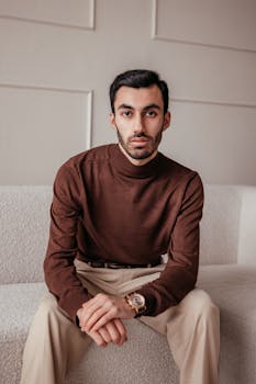 Stylish man sitting on a sofa wearing a brown sweater and beige pants indoors.
