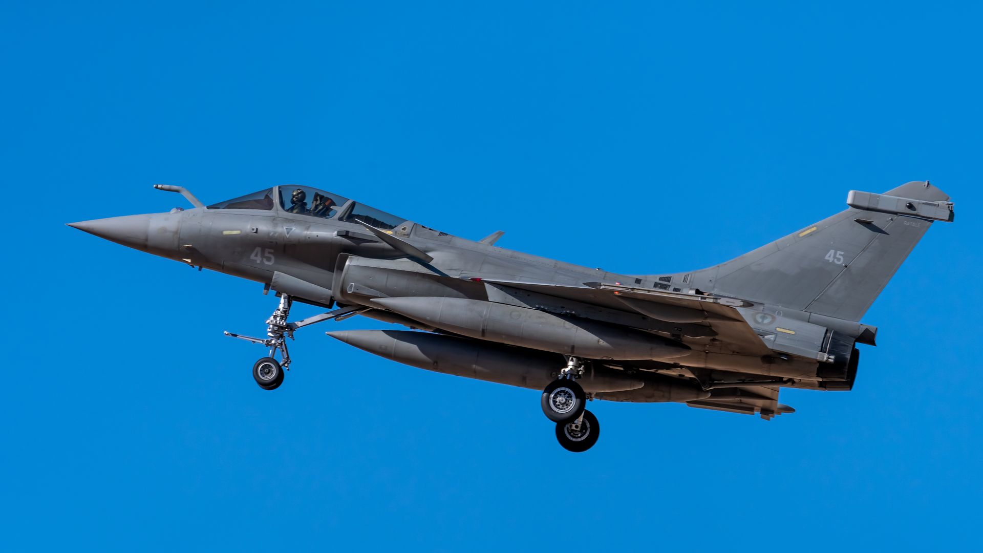 A Dassault Rafale jet flying with landing gear down against a clear blue sky in Los Llanos, Spain.