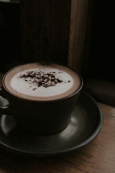 A warm cappuccino with foam in a dark ceramic cup and saucer on a wooden table.