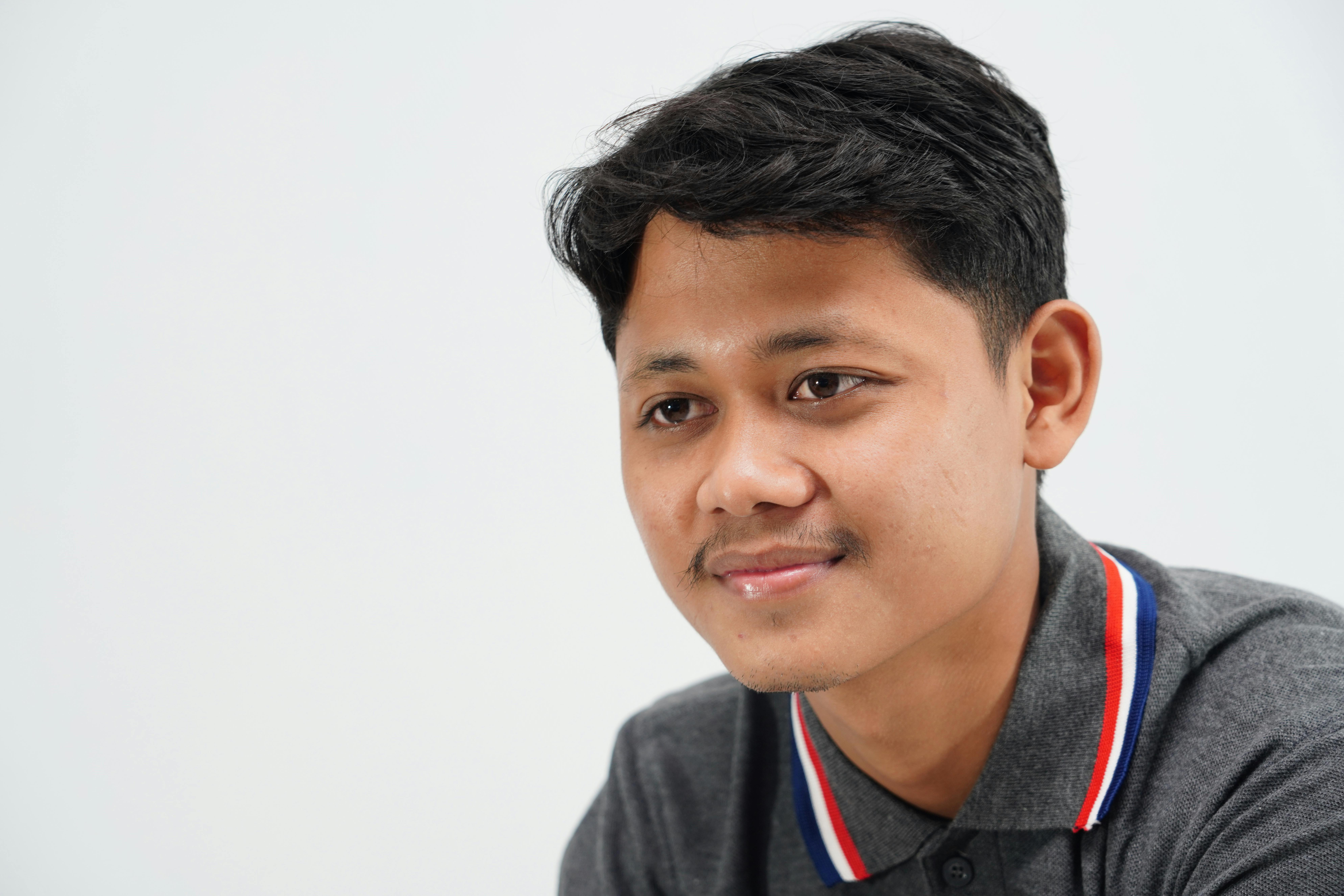 A close-up portrait of a young man smiling, wearing a gray polo shirt with a white background.