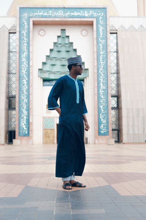 Free Man in traditional blue attire poses before architectural arch with Islamic script. Stock Photo