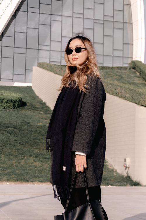 Free Fashionable woman wearing sunglasses and a gray coat stands confidently outdoors with a modern building backdrop. Stock Photo
