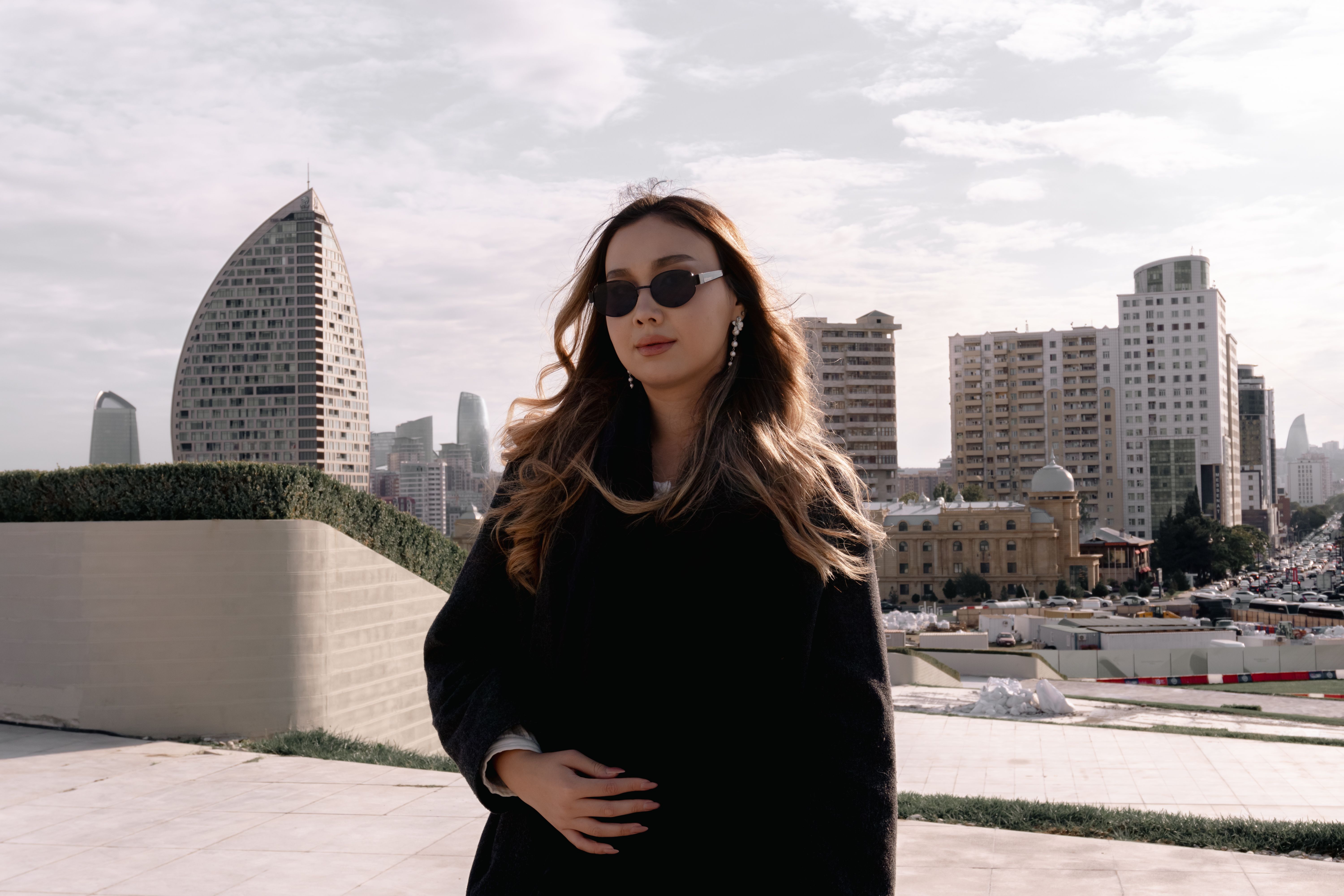Woman in sunglasses poses in front of modern city skyline with distinctive architecture, creating a trendy urban look.