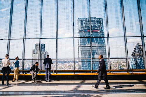 Man Walking on Sidewalk Near People Standing and Sitting Beside Curtain-wall Building