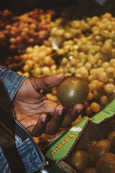 Person holding a ripe kiwi fruit at a bustling fruit market. Variety of grapes in the background.