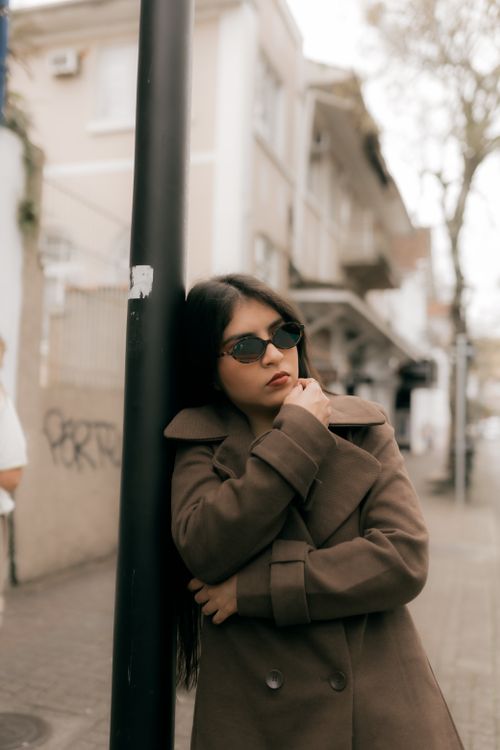 Free Fashionable woman in coat and sunglasses leaning on a pole in an urban setting. Stock Photo
