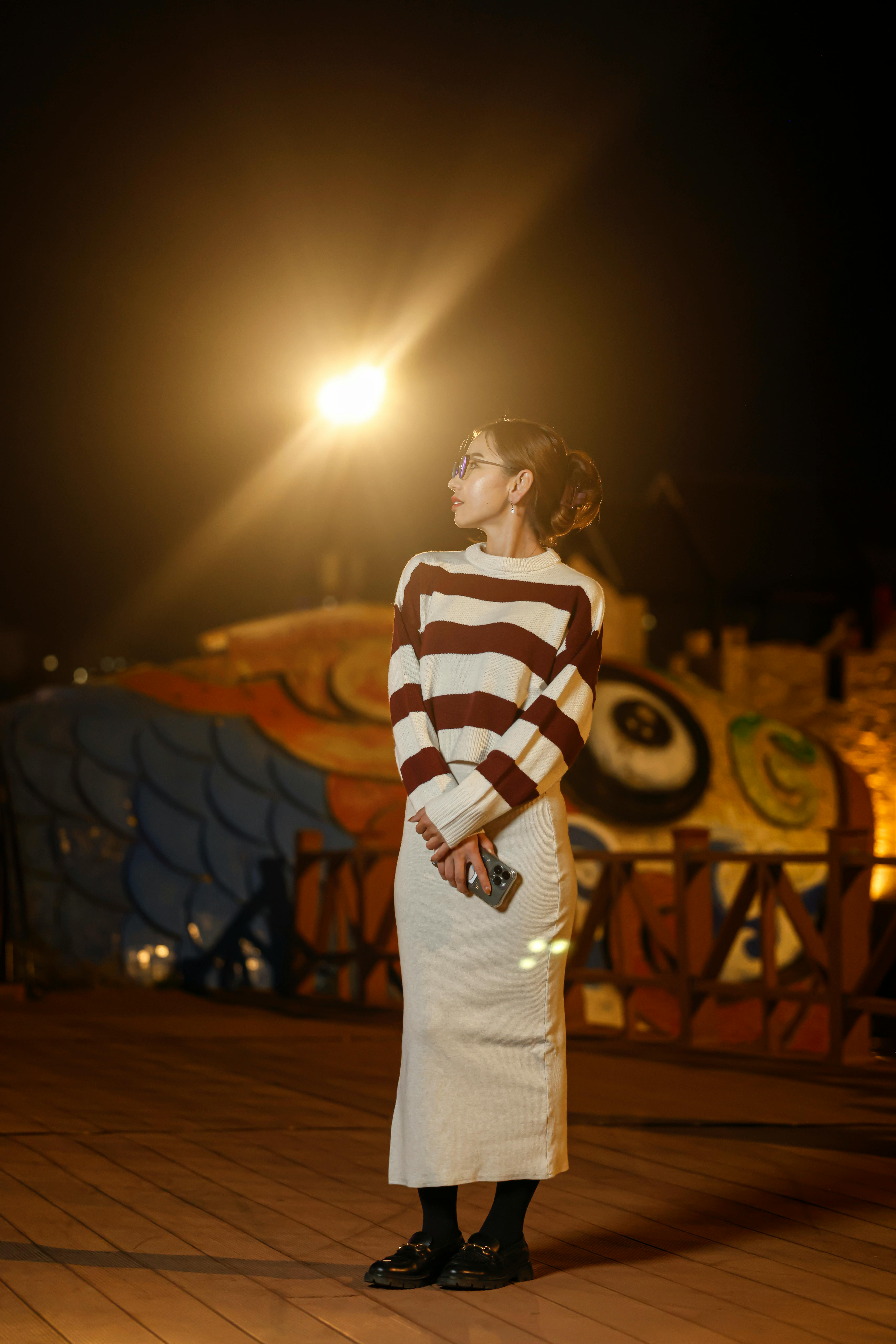 A woman stands on a decorated boardwalk at night, holding a smartphone.