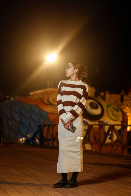 A woman stands on a decorated boardwalk at night, holding a smartphone.