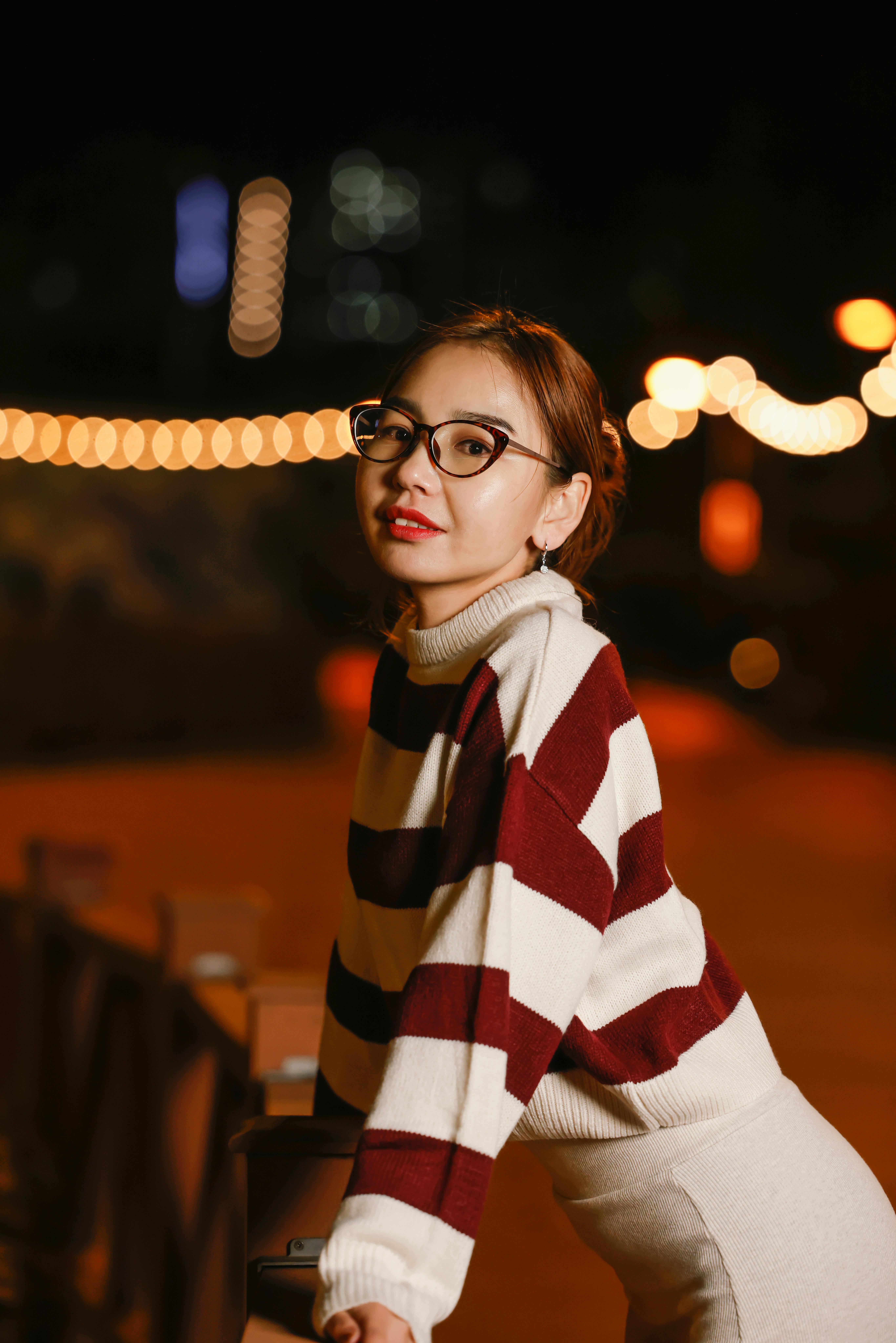 Young woman in glasses wearing a striped sweater, posing outdoors at night.