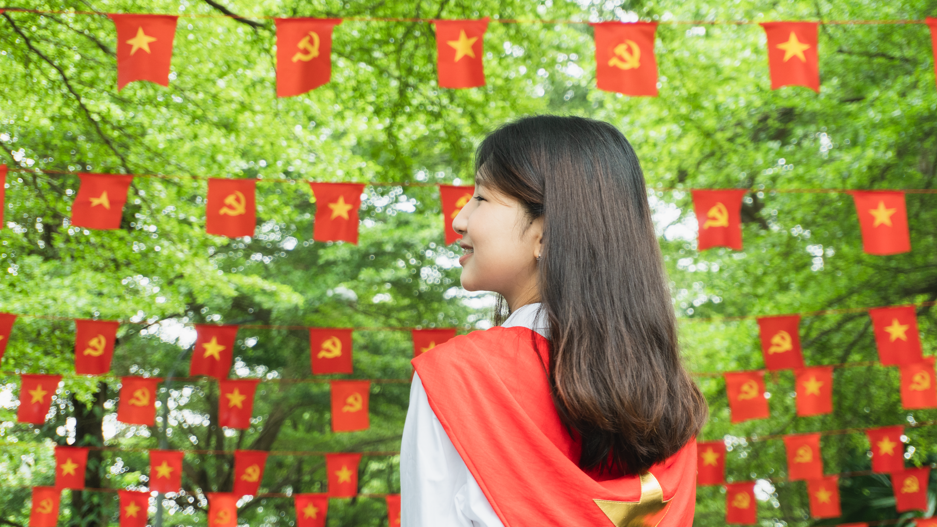 Smiling woman in red vest outdoors surrounded by red flags with stars and symbols.