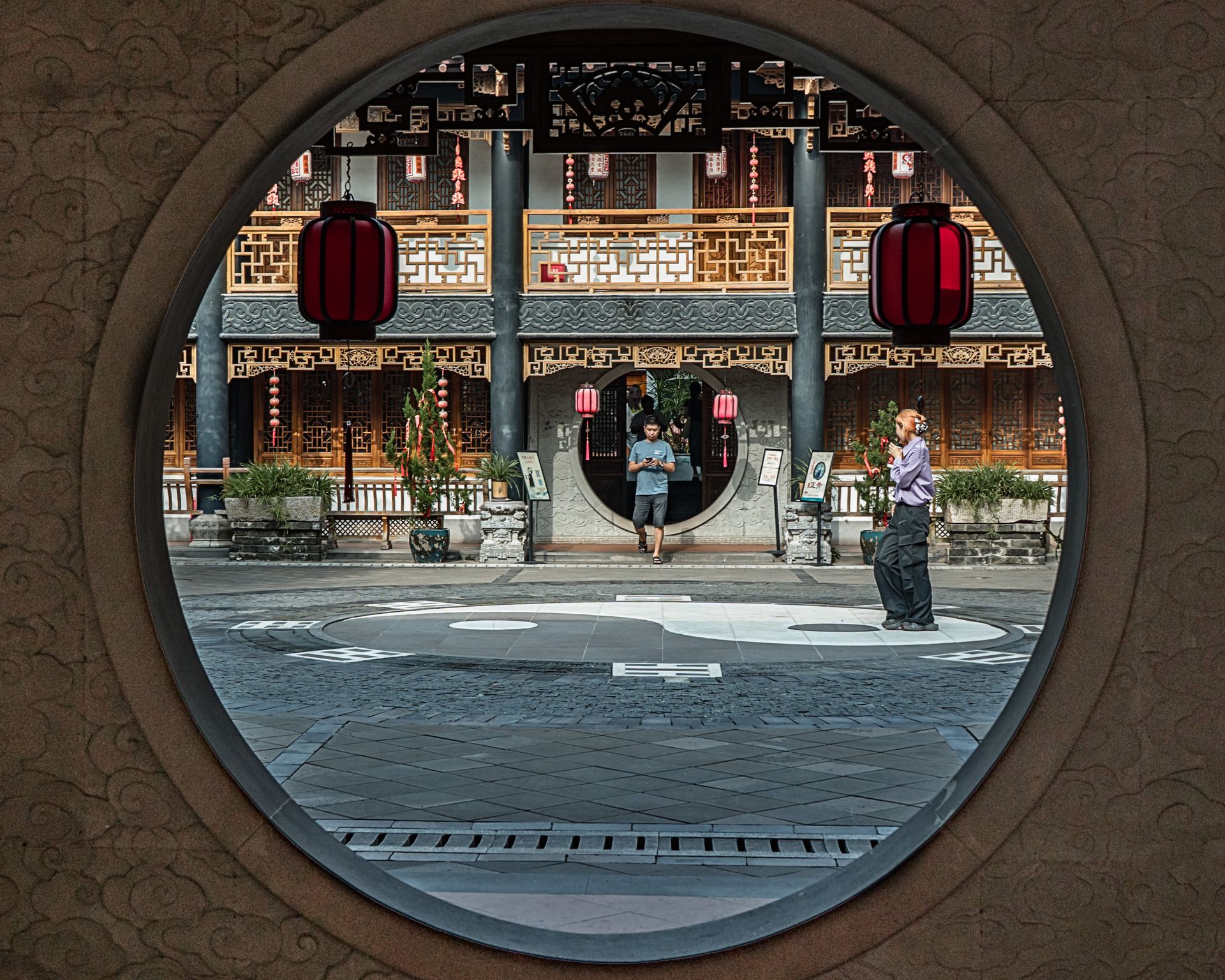 View through a circular doorway to a courtyard with traditional Chinese architecture and red lanterns.