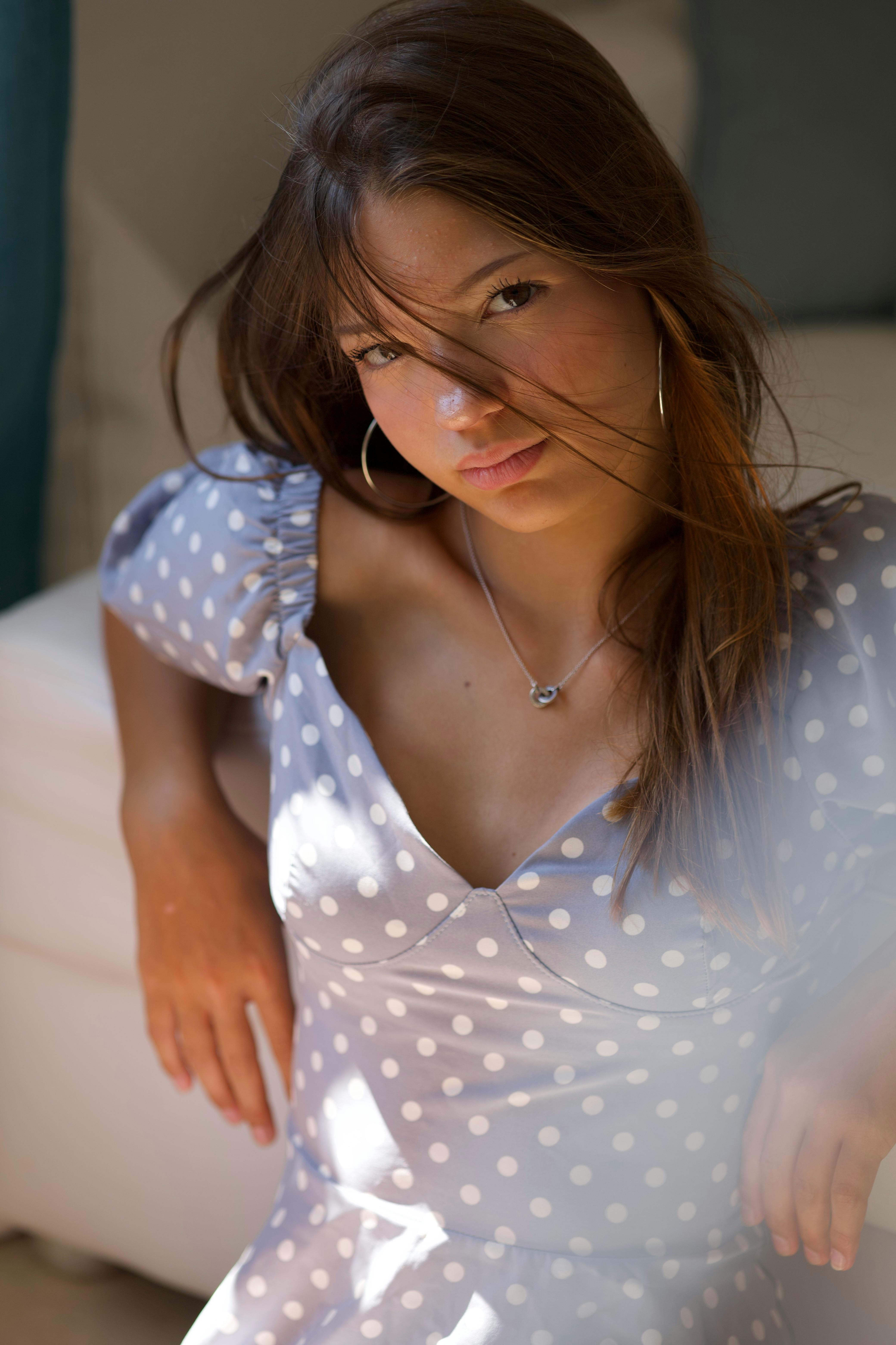 Portrait of a young woman wearing a polka dot dress, sitting indoors with soft lighting and a relaxed pose.
