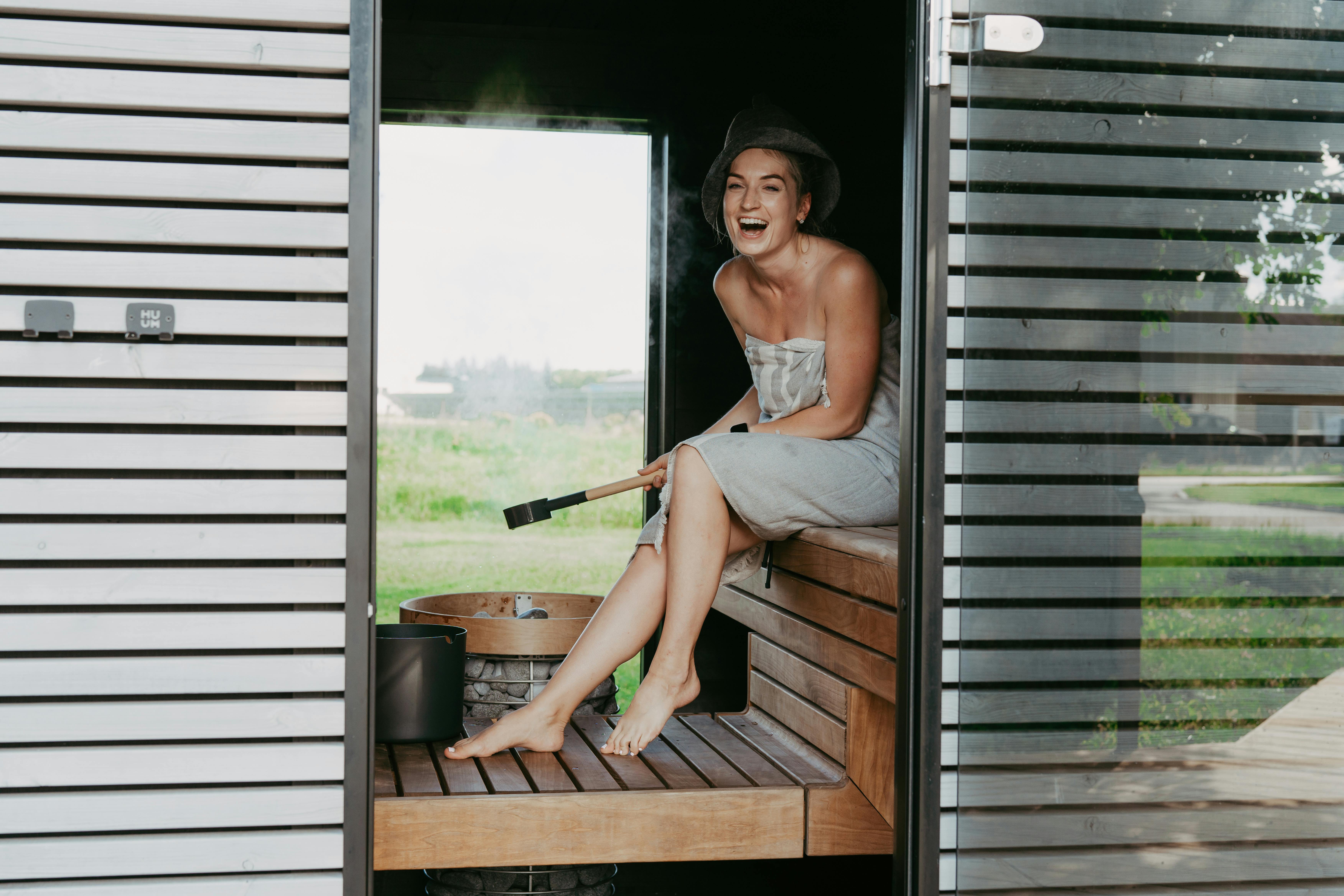 Une femme riant et se relaxant dans un sauna moderne avec un chapeau de sauna, profitant de la vapeur et de la chaleur.