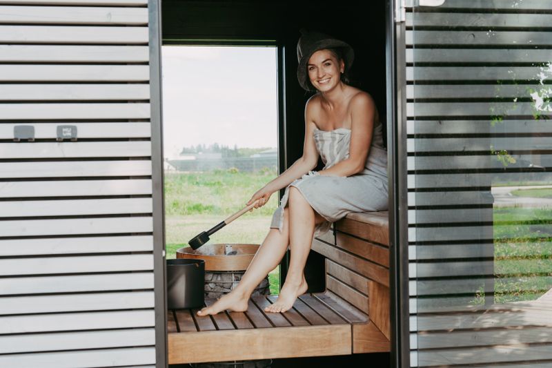Woman in outdoor traditional sauna pouring water on hot stones with ladle