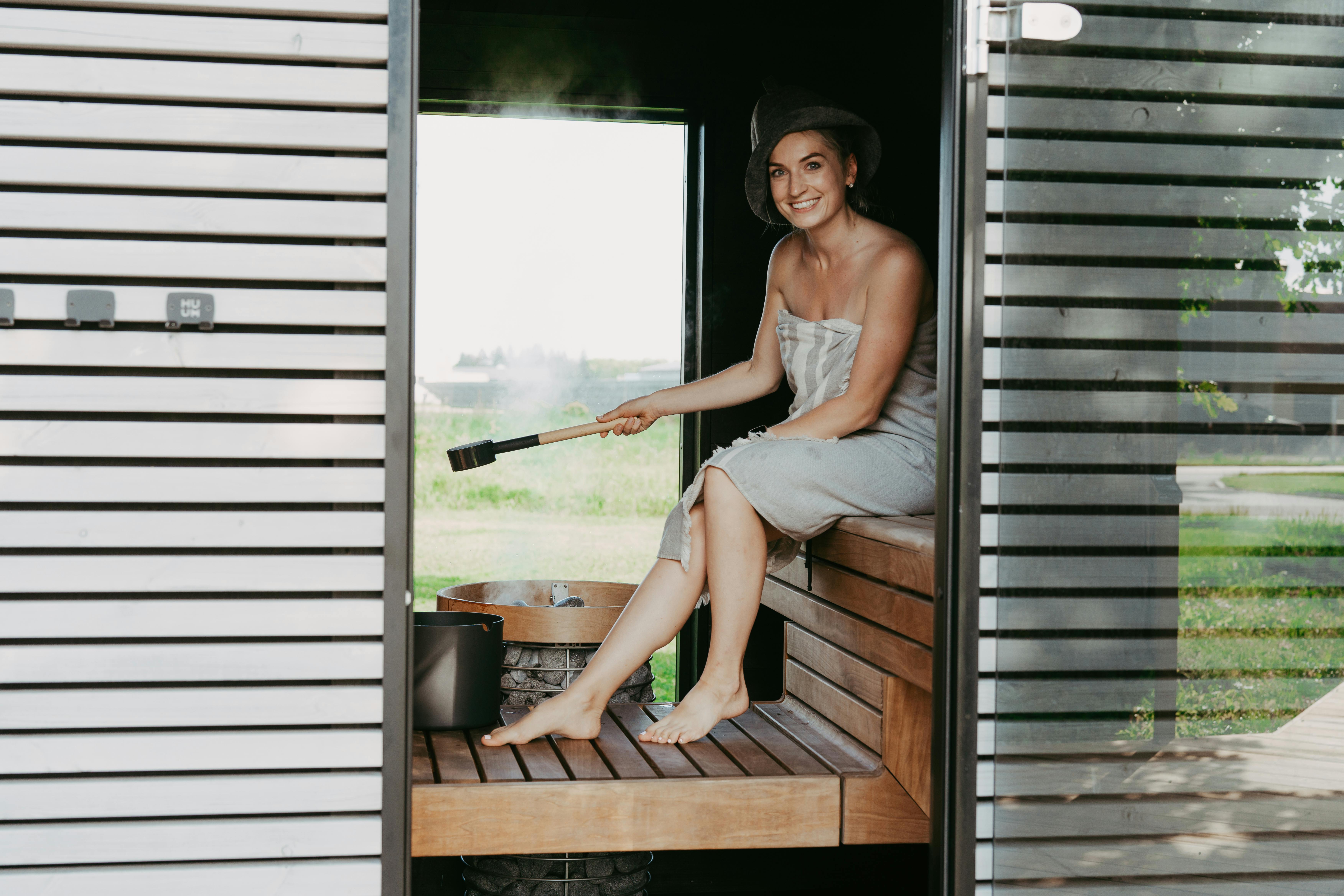 Femme adulte dans un sauna portant une serviette et un chapeau, assise sur un banc, profitant d'un moment de détente.