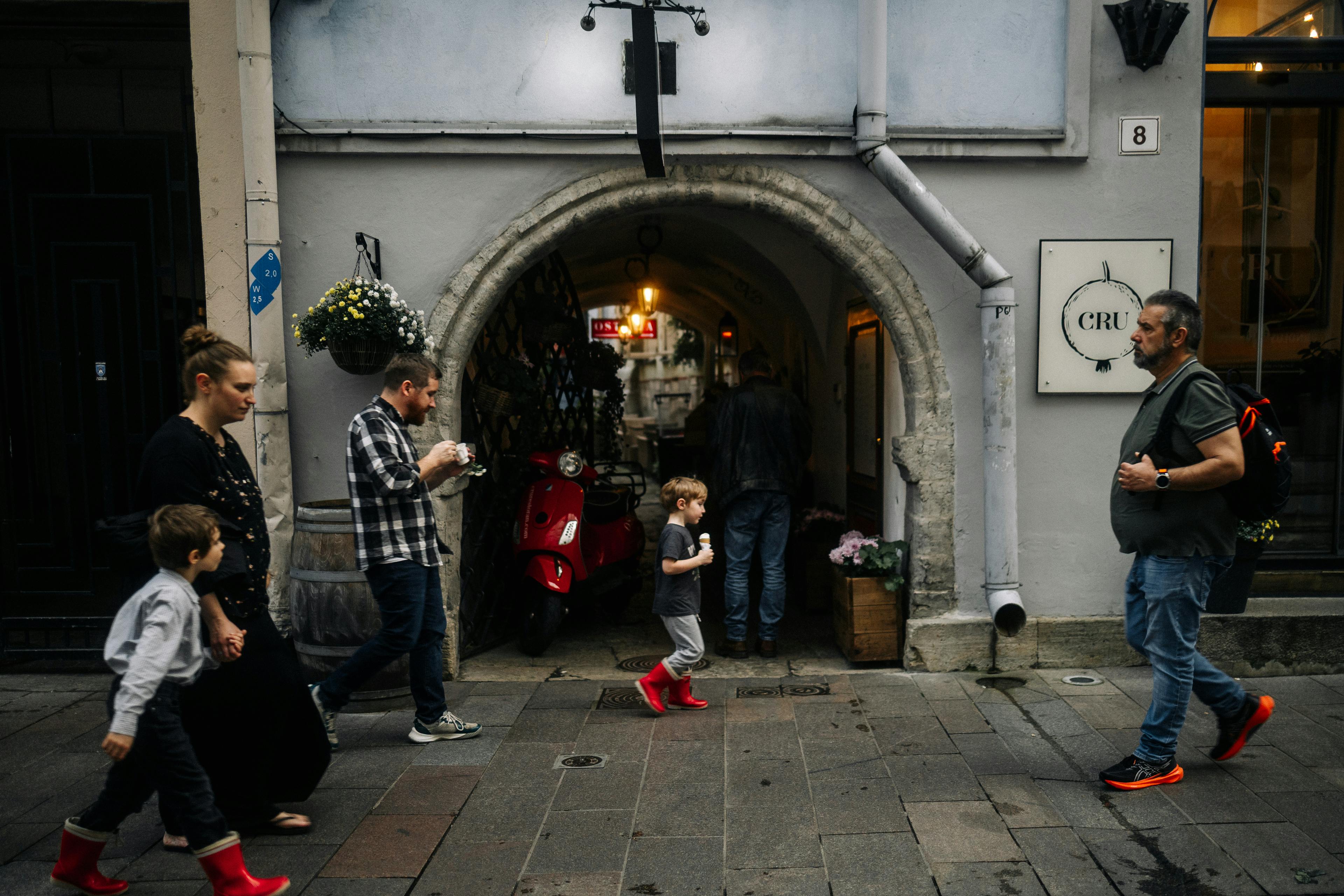 A lively street scene with pedestrians walking past a vintage archway featuring red scooter and hanging flowers.