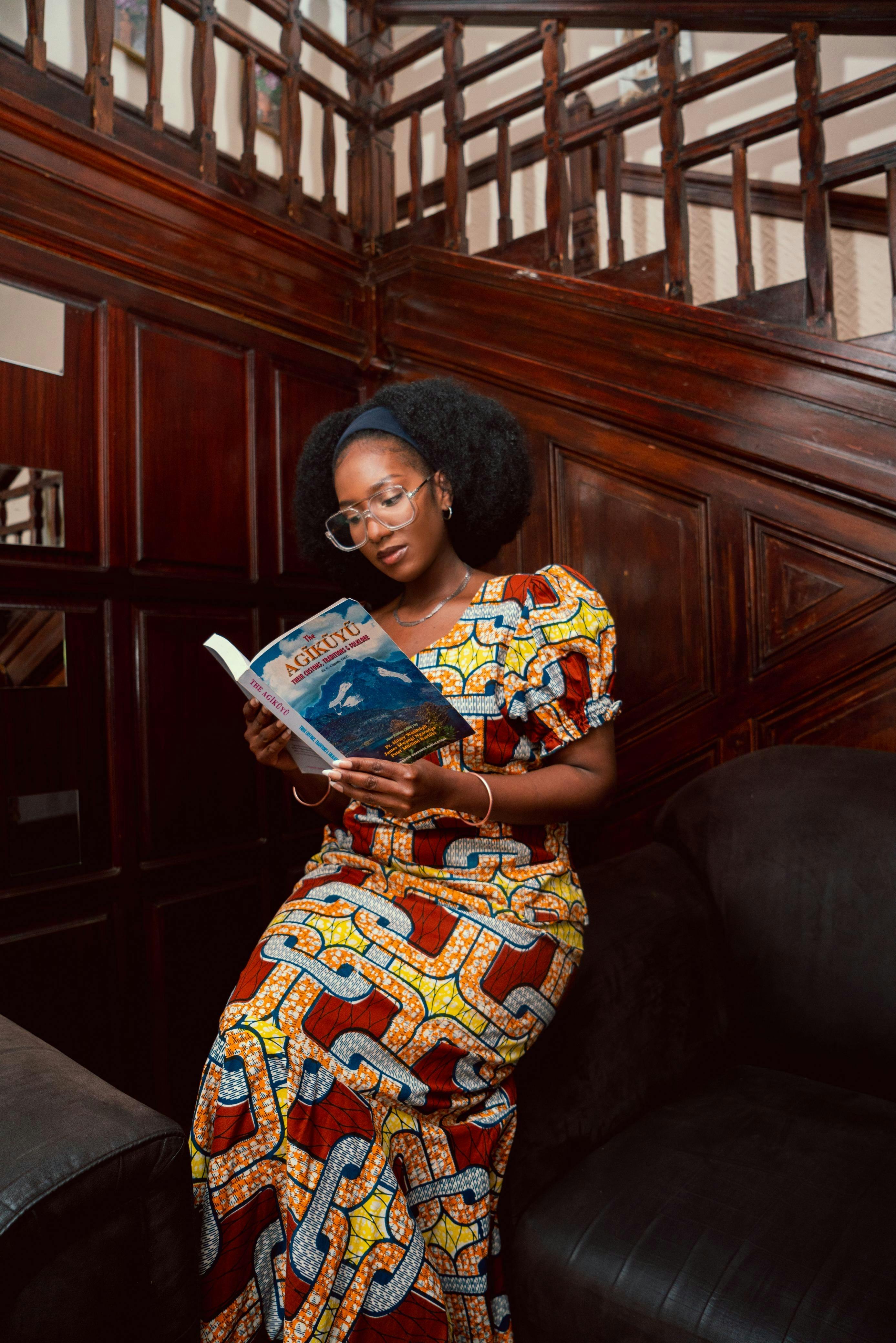 A woman in traditional African attire reading a book indoors, with rich wood paneling.