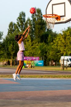 A young girl in mid-jump shooting a basketball into a hoop in an outdoor park setting.