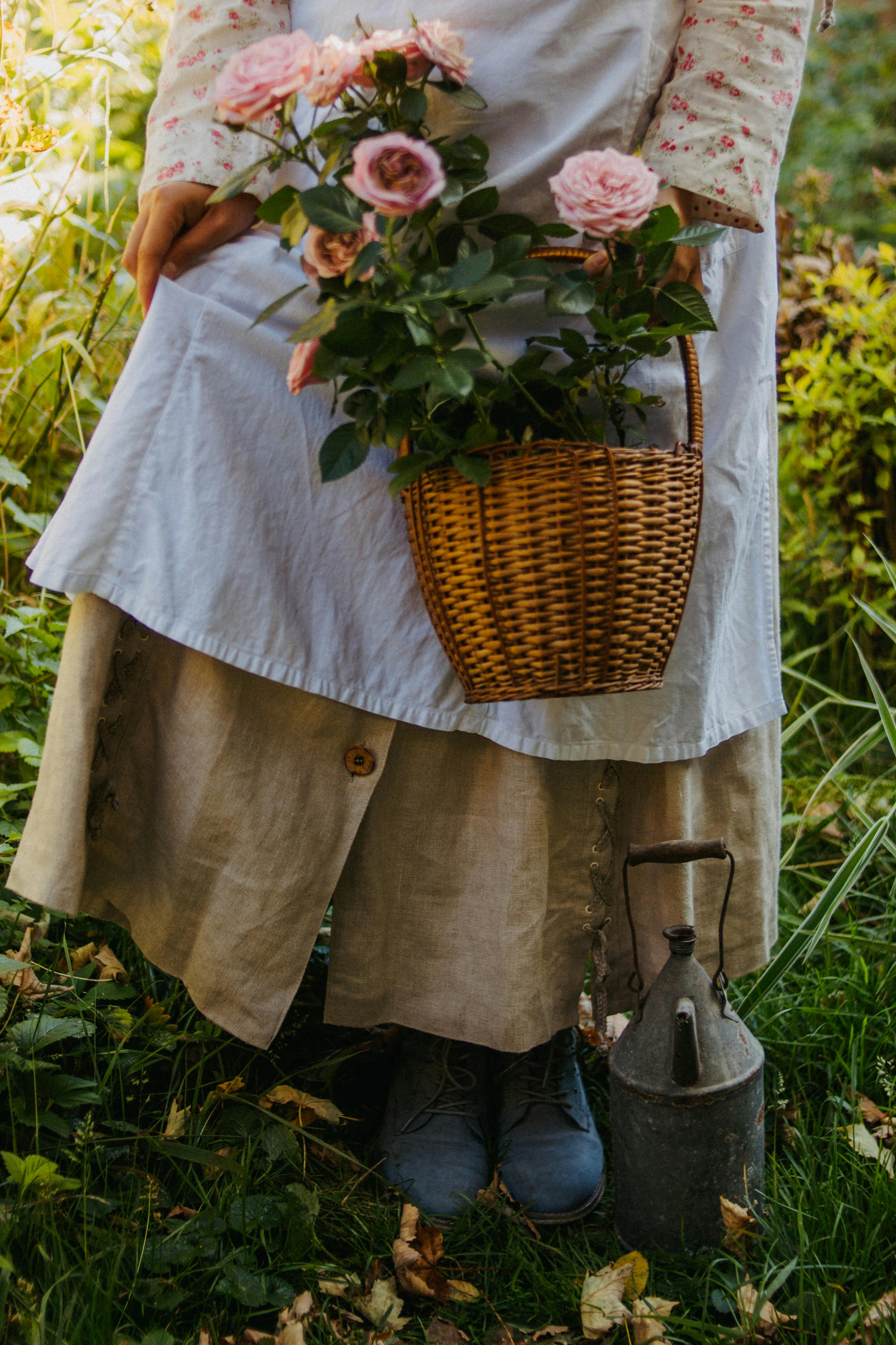 A woman in a vintage dress holds a basket of pink roses in a lush garden setting.