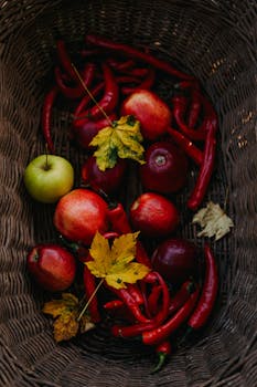 A wicker basket filled with red apples, green apple, and red chili peppers, adorned with autumn leaves.