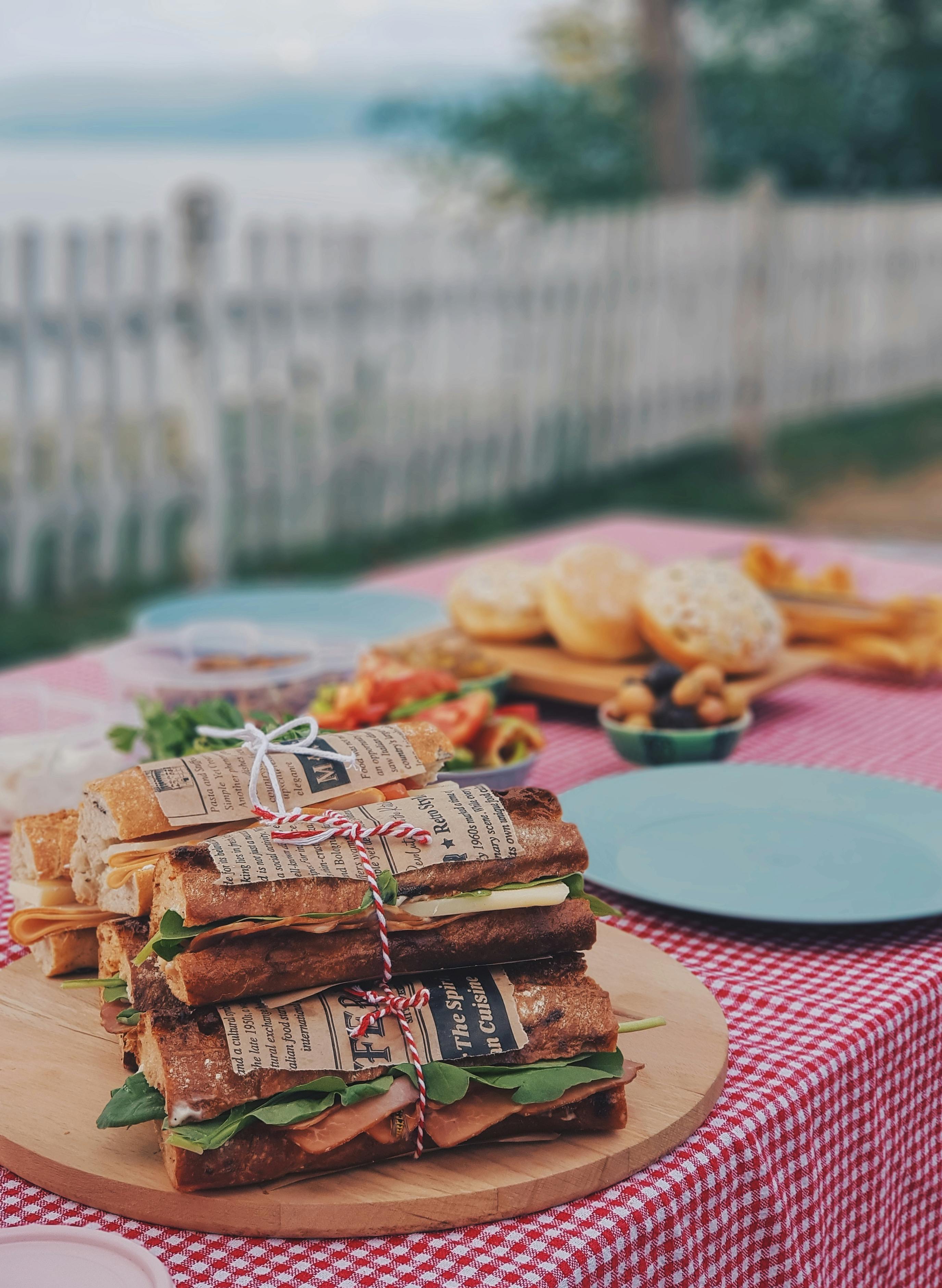 Gourmet sandwiches with fresh greens and meats on a picnic table by the lake.