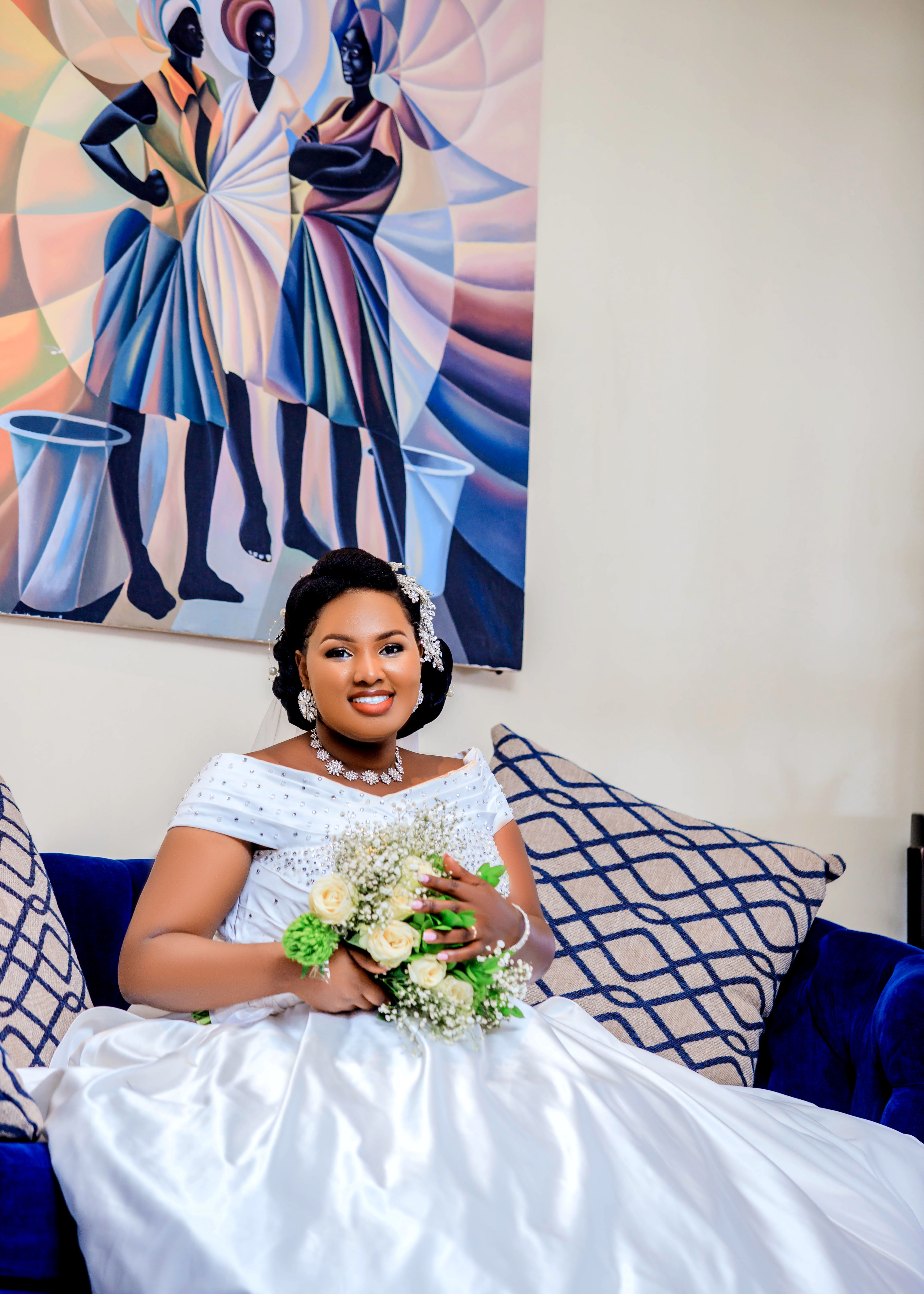 Elegant bride in a white gown holding a bouquet, sitting on a sofa indoors.