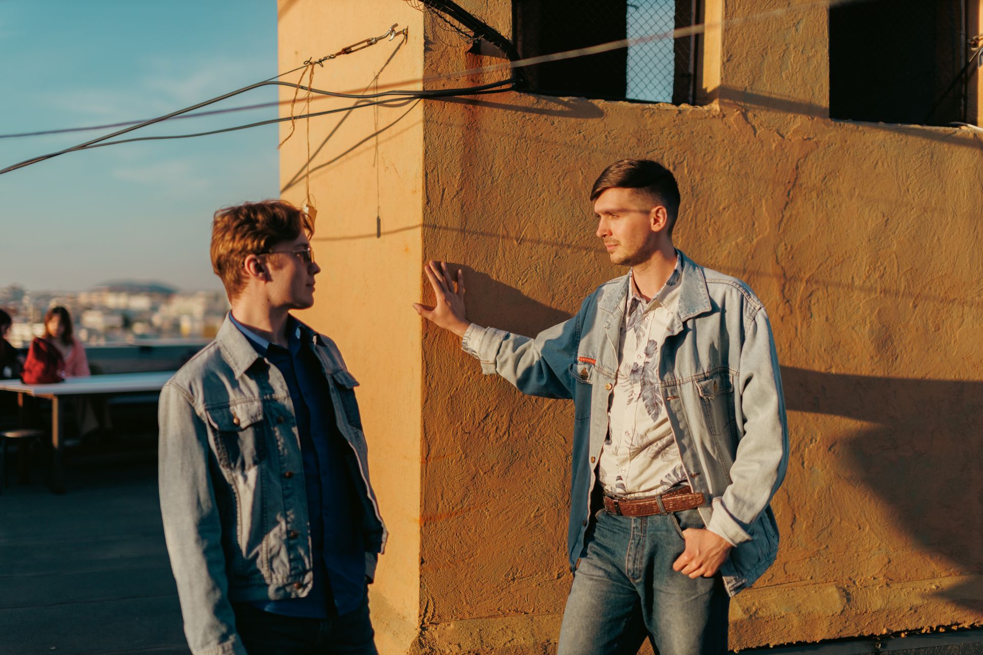 Two men wearing denim jackets engage in a casual conversation on a sunlit rooftop.