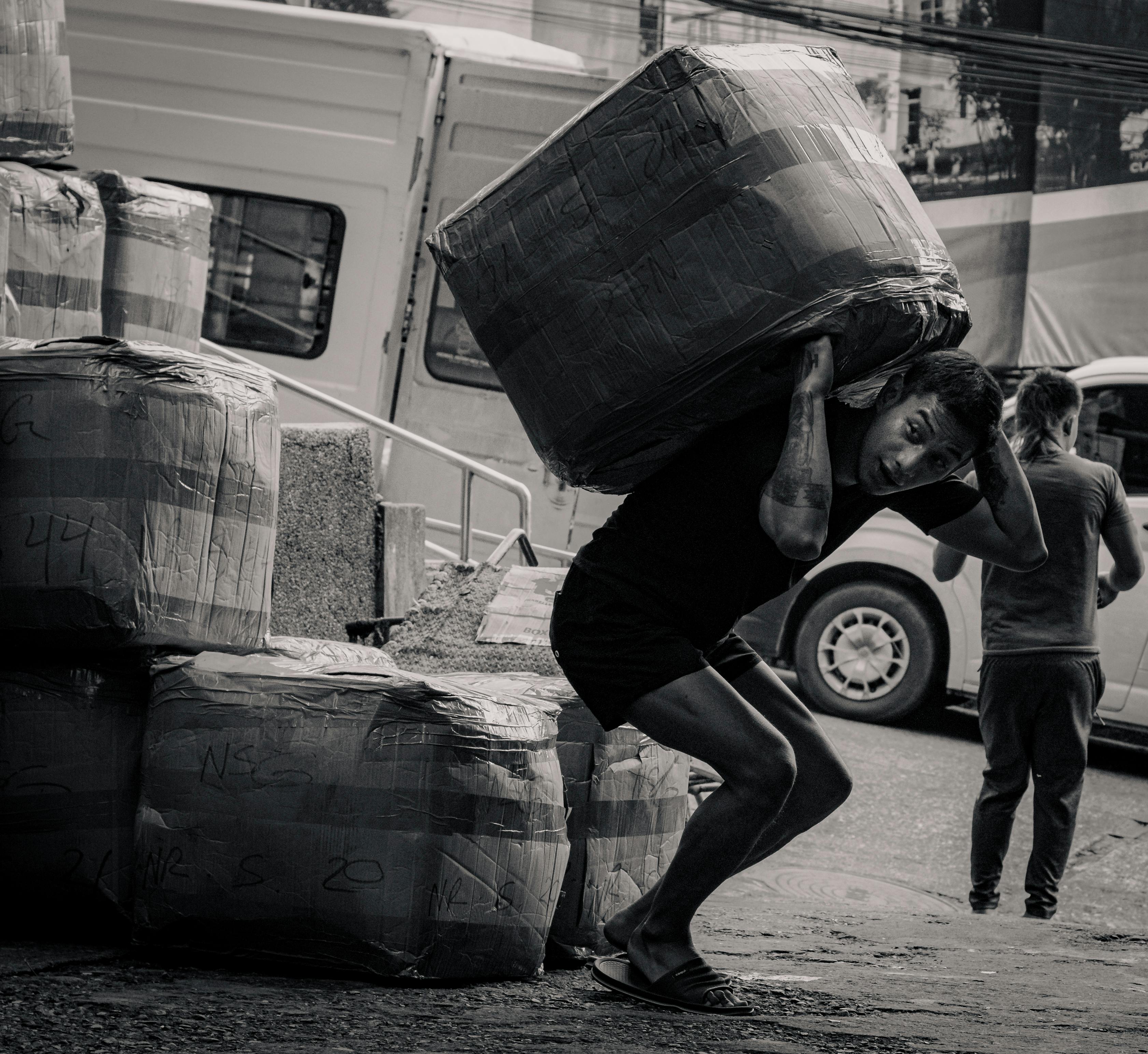 A black and white photo capturing a porter carrying a large package at a market in Baguio, Philippines.