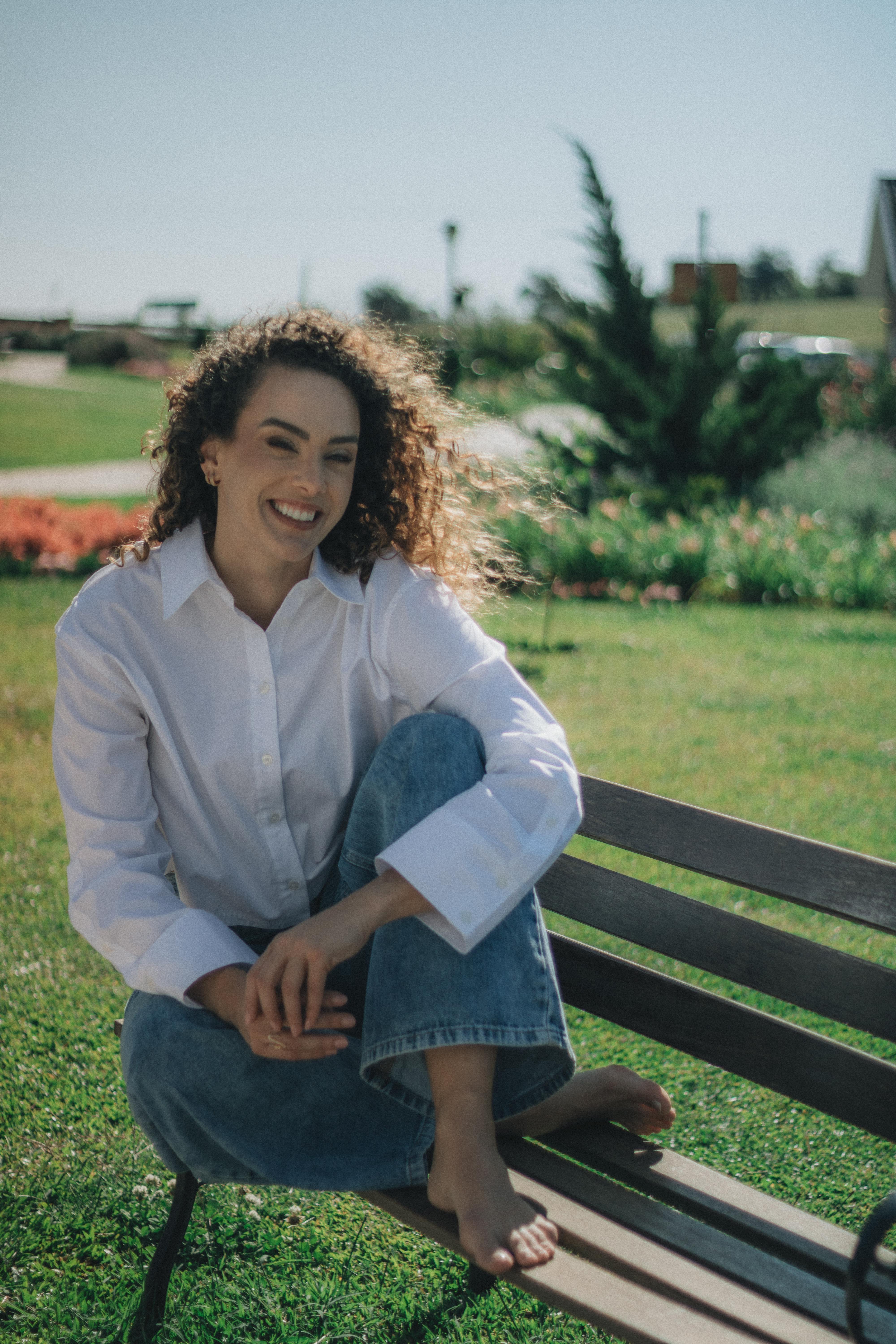 Безкоштовне Життєрадісна жінка в повсякденному одязі насолоджується сонячним днем ​​на лавці в парку, оточена зеленню. стокове фото