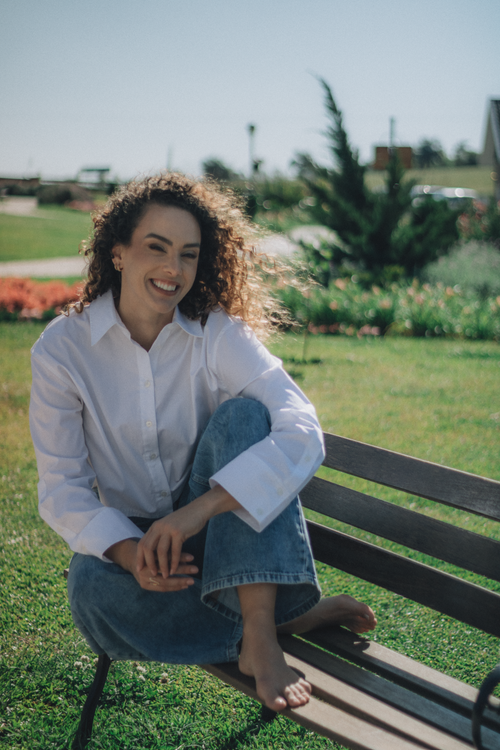 Free A cheerful woman in casual attire enjoys a sunny day on a park bench, surrounded by greenery. Stock Photo