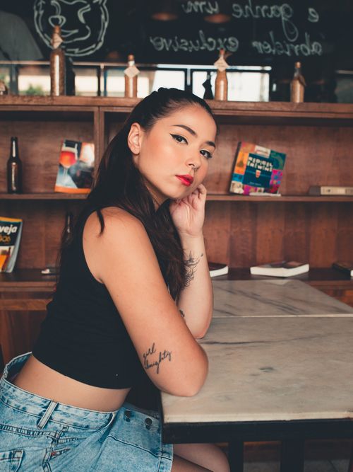 Free Young woman in casual outfit posing at a cafe table with tattoos and a confident expression. Stock Photo