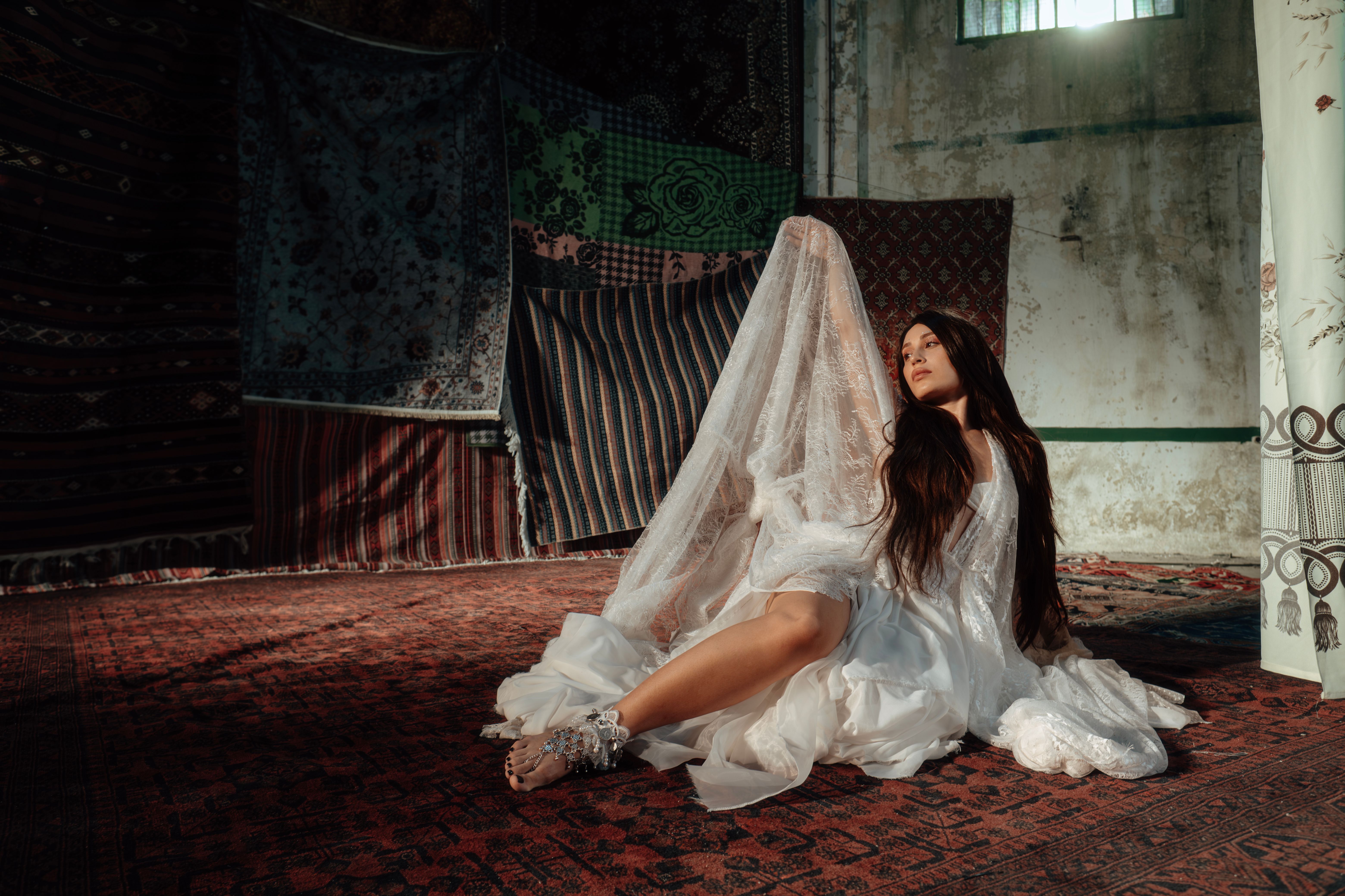 Woman in flowing white dress poses dramatically on a carpeted floor, surrounded by patterned textiles.