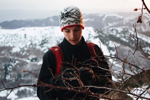 一名穿着冬装的年轻人在雪山背景下检查贫瘠树枝上的相机