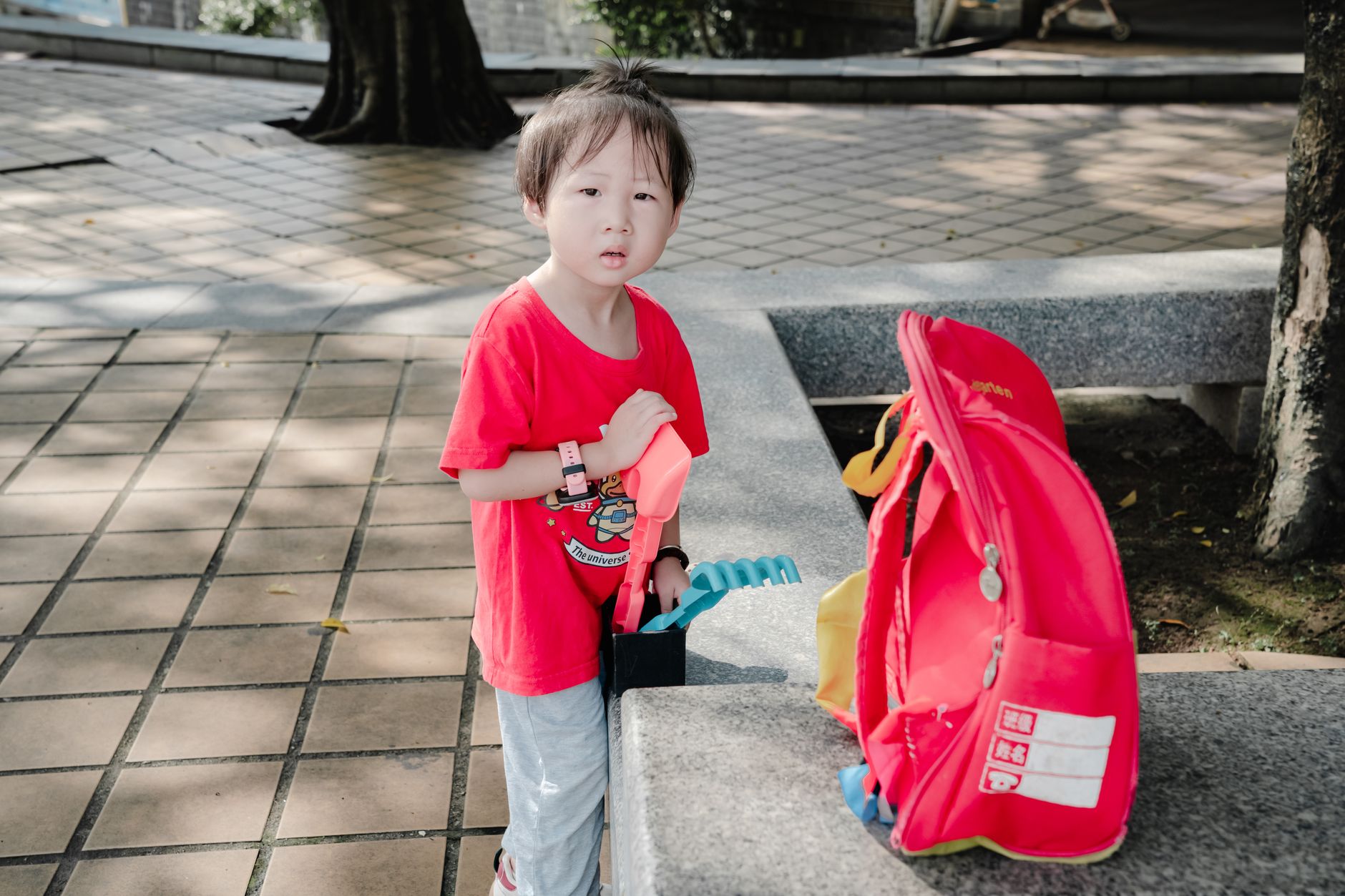 A young child standing outdoors with a backpack in a sunny park setting.