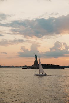 A sailboat glides by the iconic Statue of Liberty during a picturesque sunset over New York Harbor.