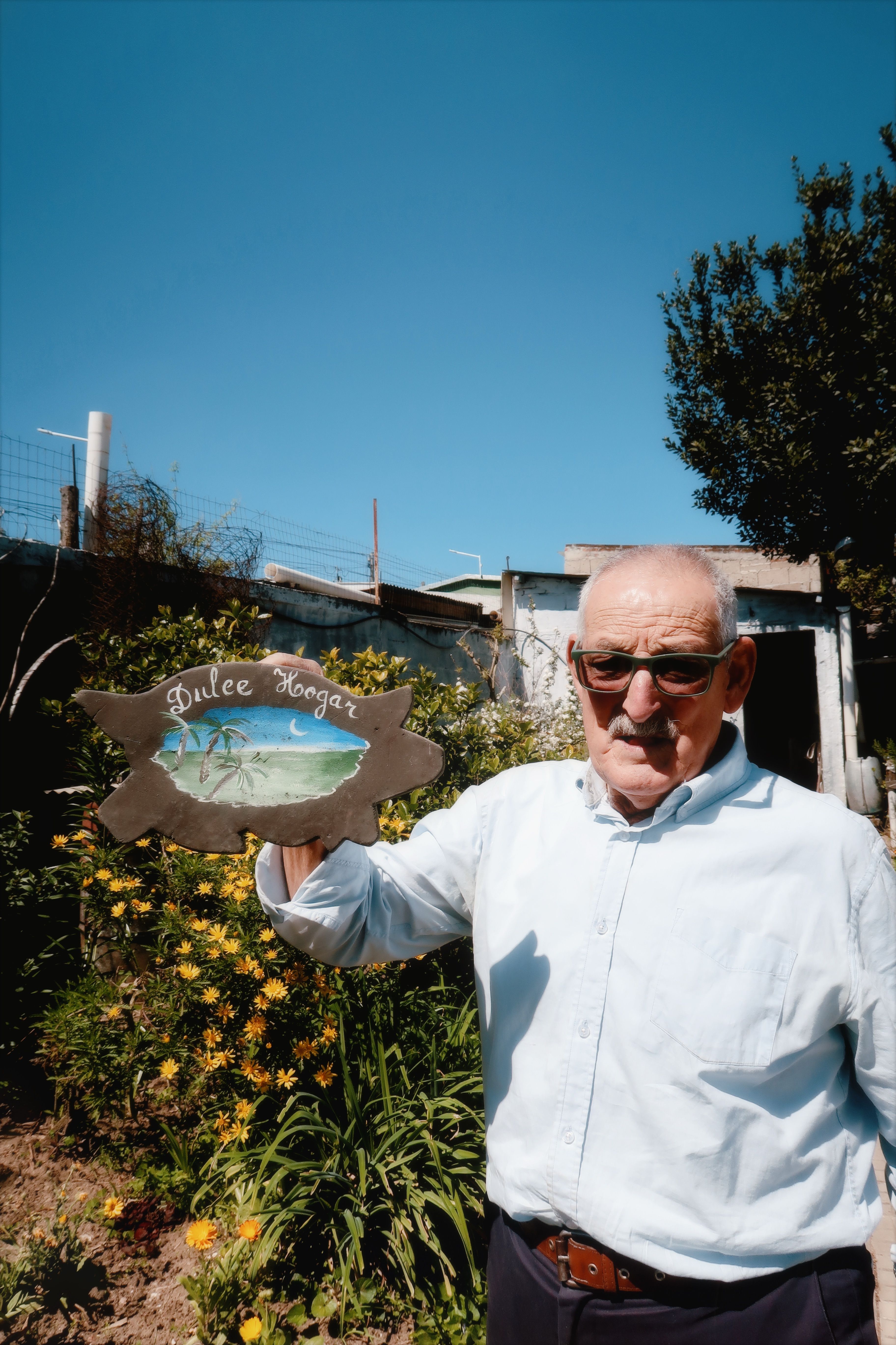 Elderly man in garden holding sign reading 'Dulce Hogar' under a clear blue sky.