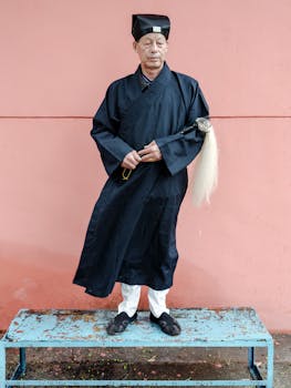 Elderly man in traditional Asian attire poses against a pink wall, displaying cultural heritage.