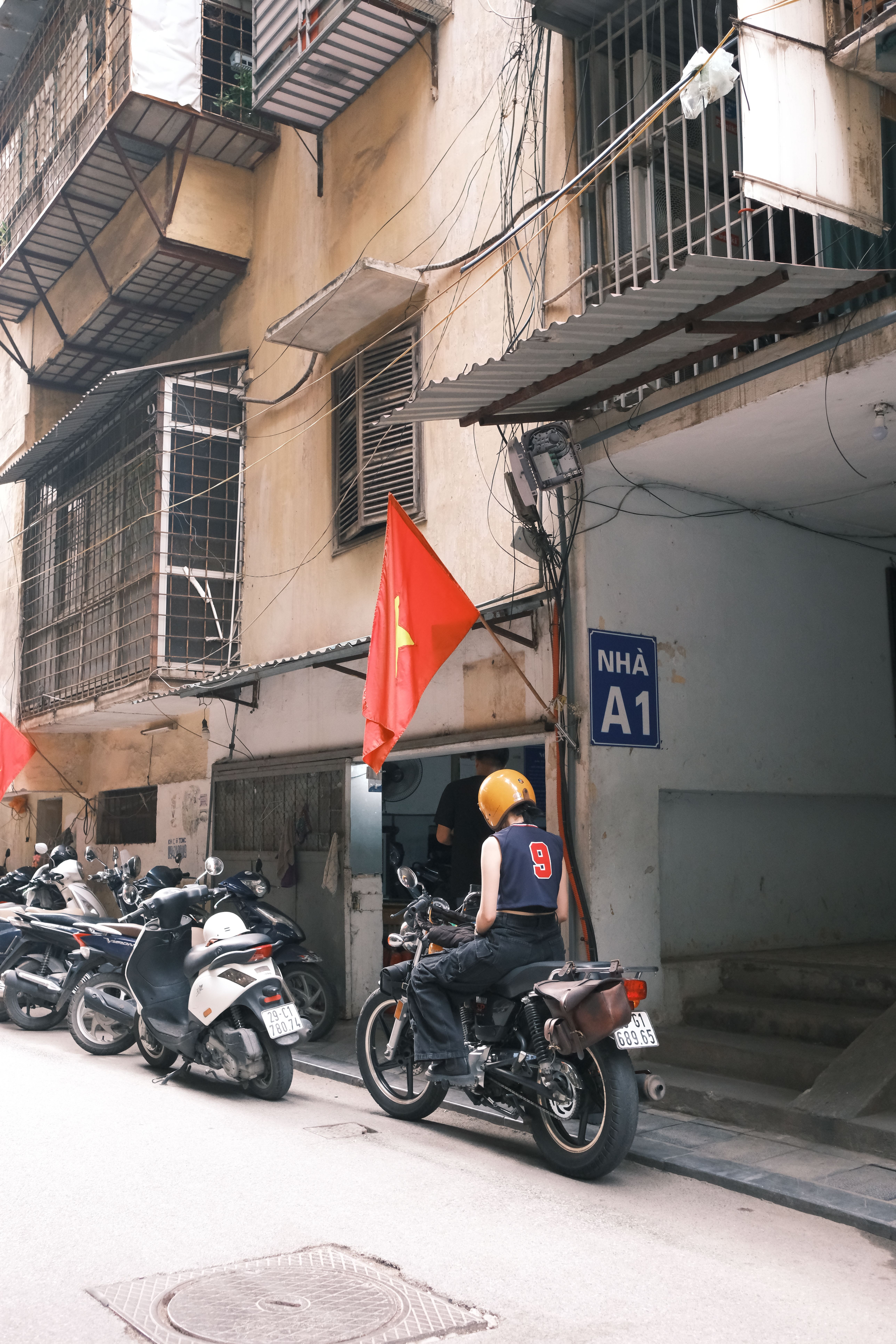 A person rides a motorcycle past scooters and buildings adorned with Vietnamese flags on a city street.