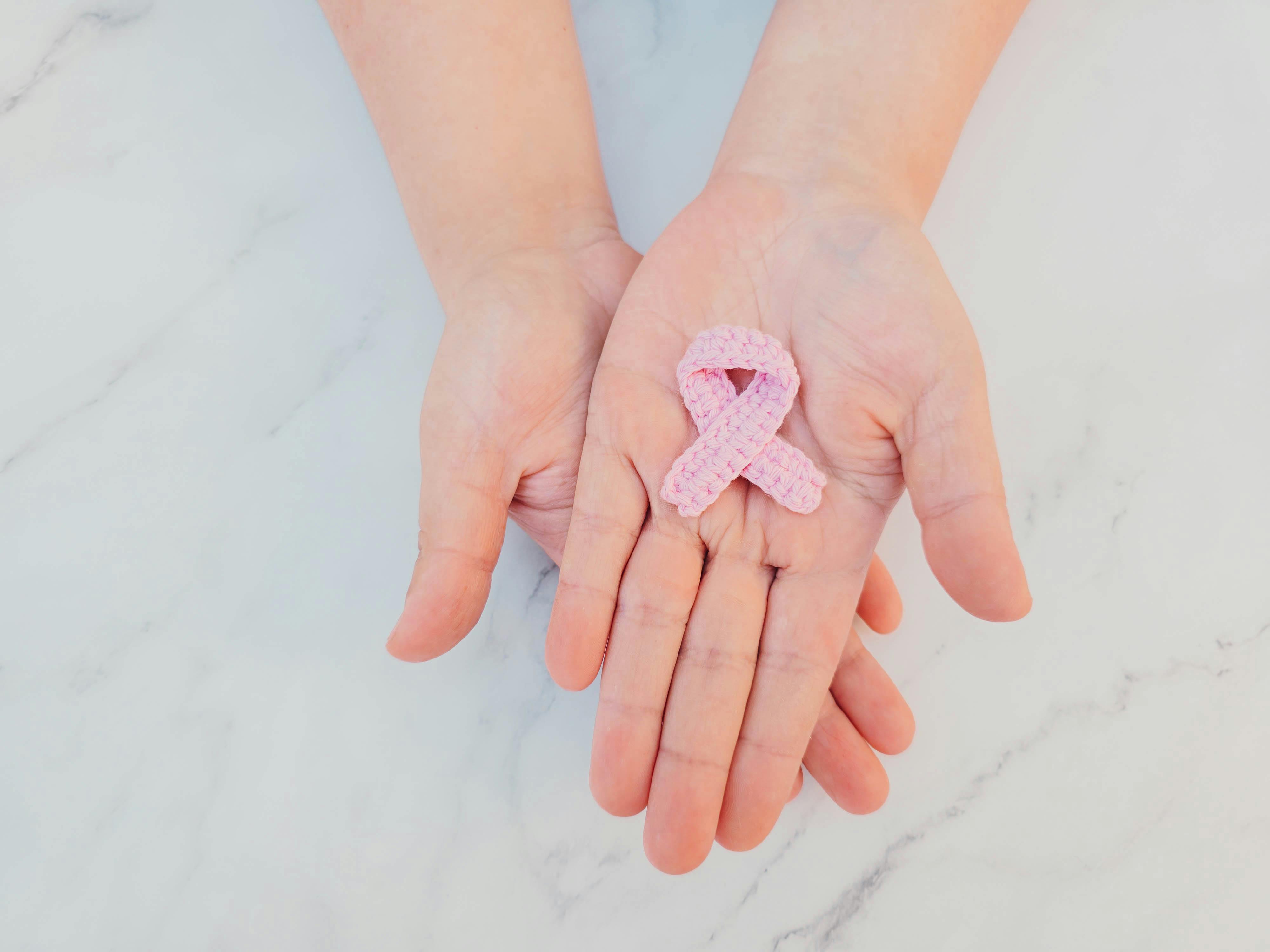 Pink ribbon in hands symbolizing breast cancer awareness on marble background.