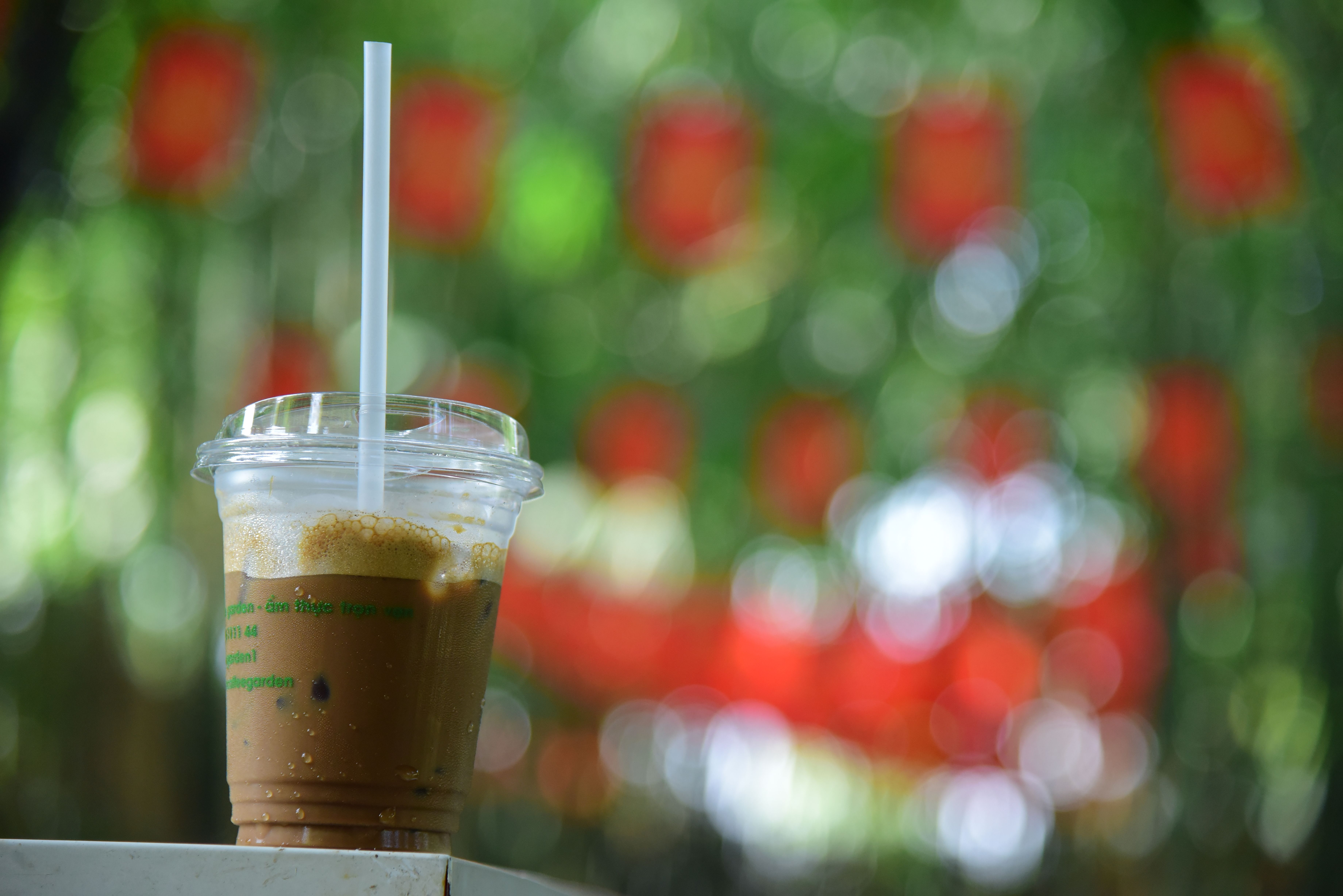 A close-up of iced coffee in a clear cup with green and red bokeh background outdoors.