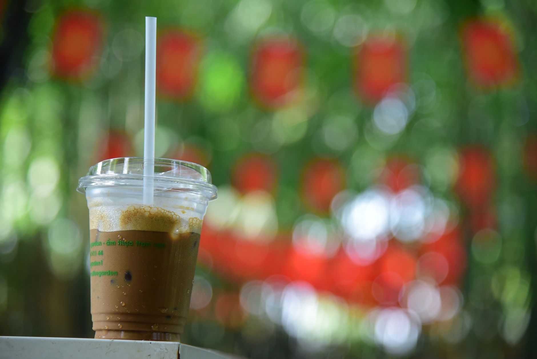 A close-up of iced coffee in a clear cup with green and red bokeh background outdoors.