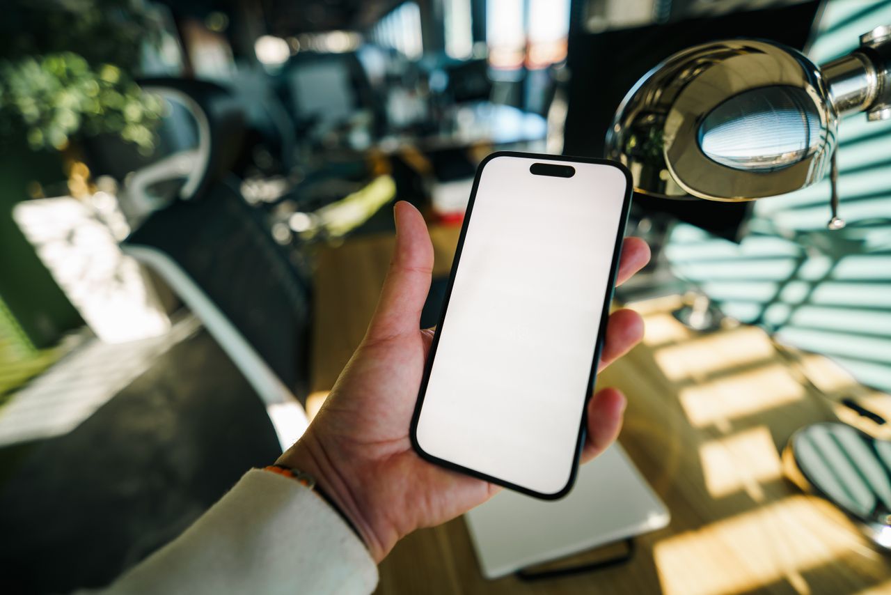 A hand holds a smartphone with a blank screen in a workspace.