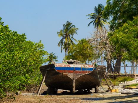 A rustic fishing boat rests on a sandy shore surrounded by palm trees and clear blue skies.