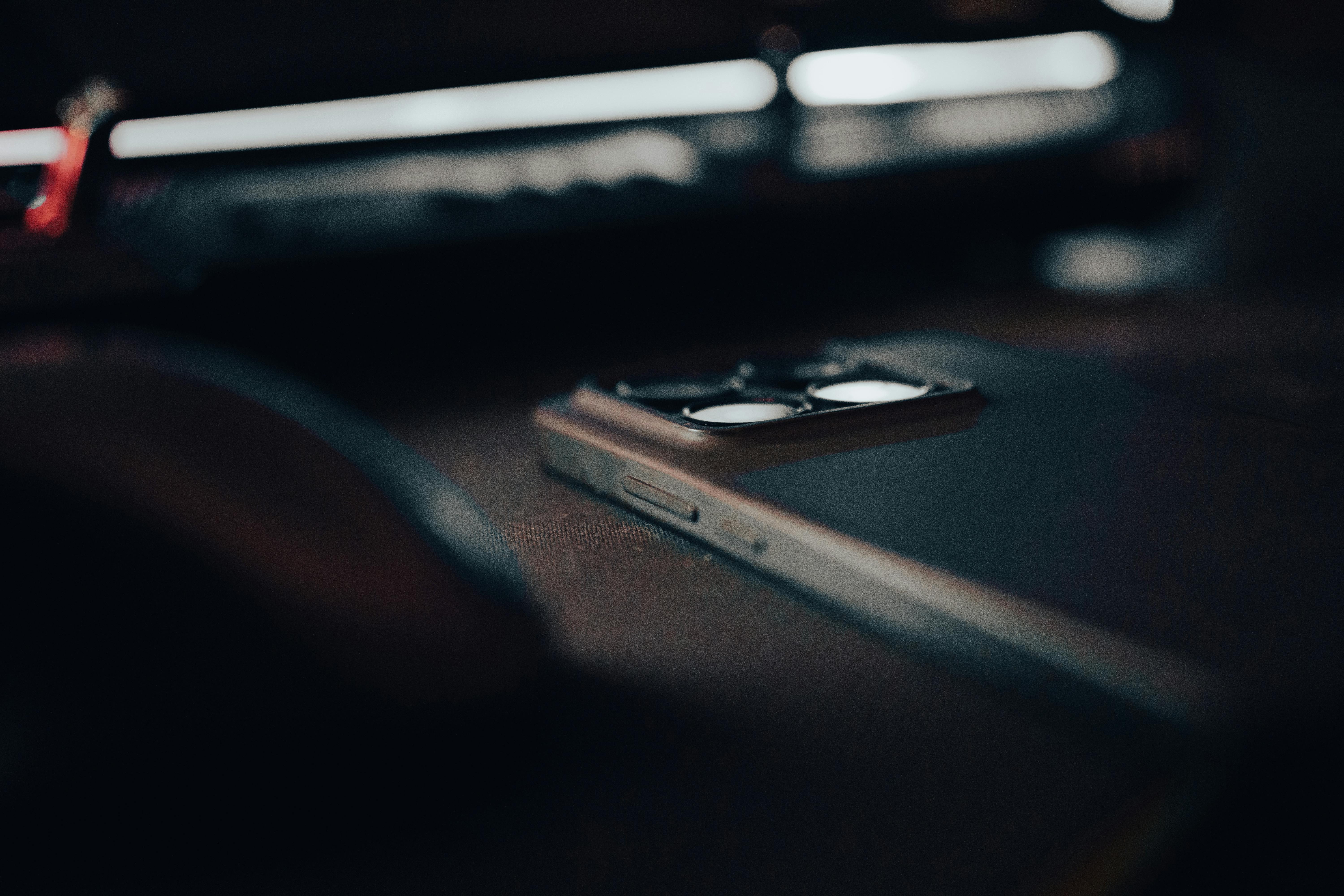 Free A sleek smartphone lies on a desk with a blurred electronic device in the background, exuding a moody tech vibe. Stock Photo