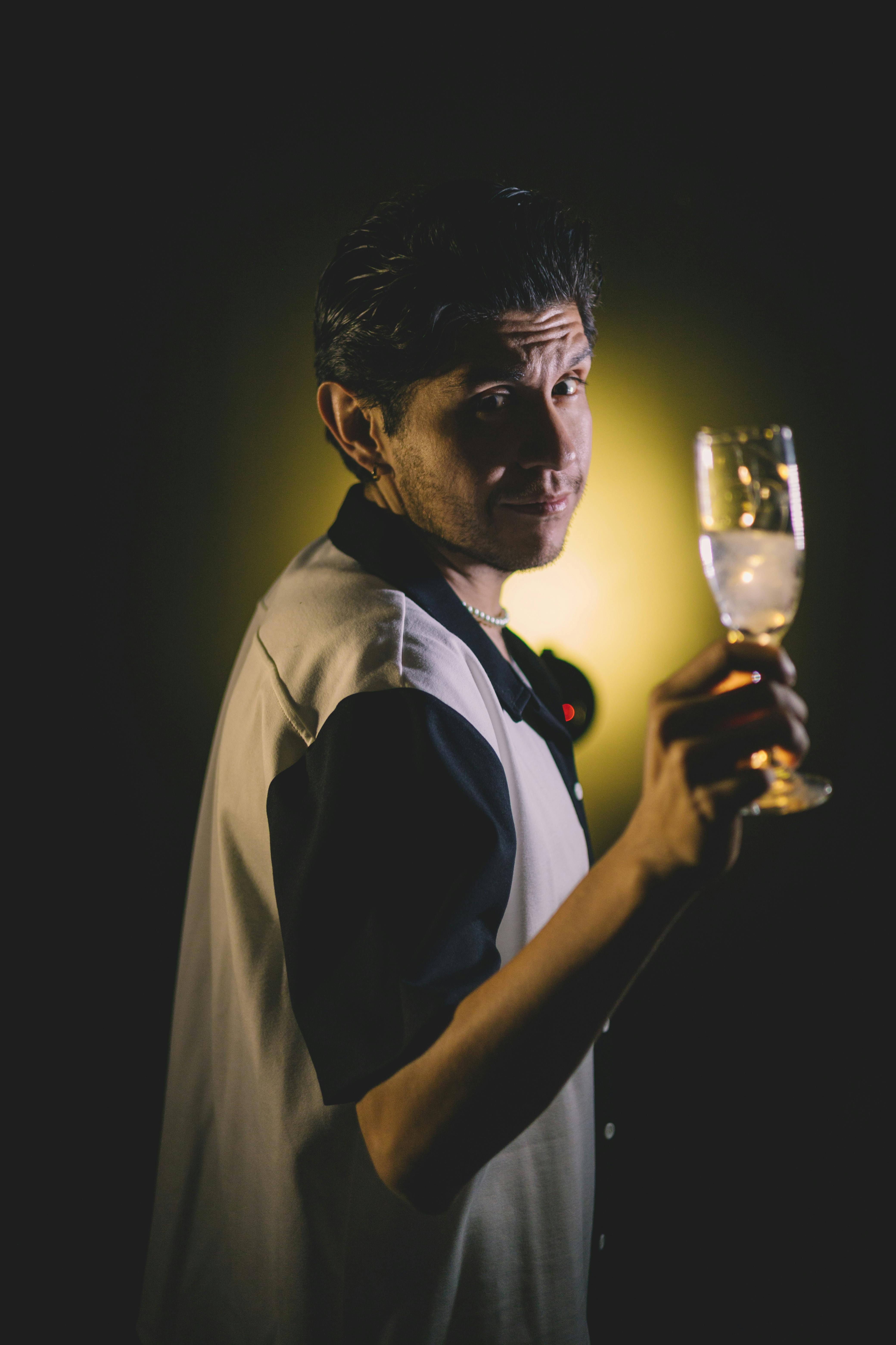 A man in casual attire holding a wine glass, highlighted against a dark background.