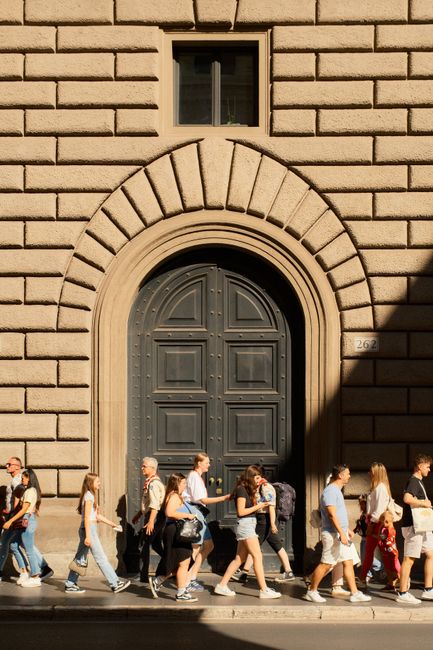 People walk past a grand stone entrance in bright daylight showcasing vibrant street life.