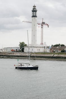 A sailboat passes a lighthouse under cloudy skies in Dunkerque, France.